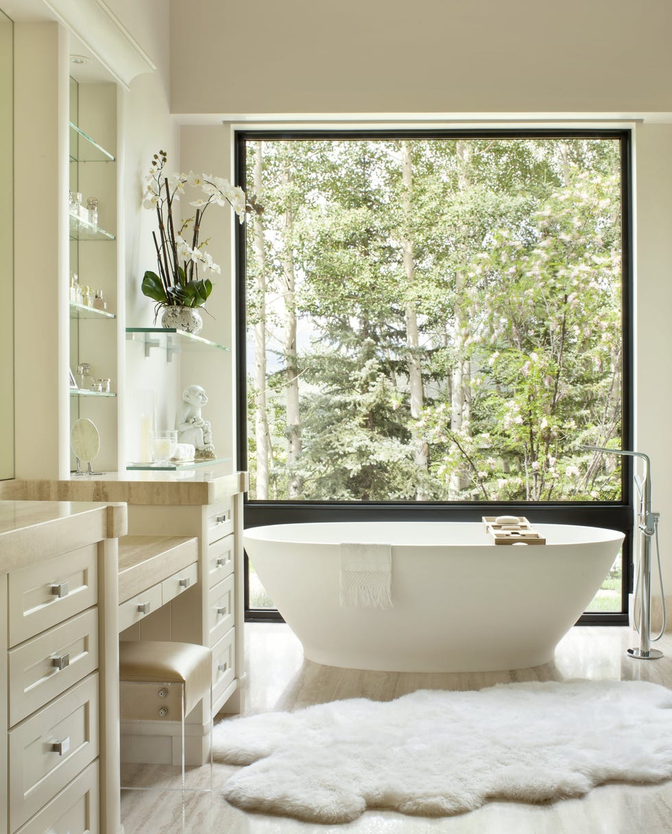 bathroom with plush white rug at the foot of the tub and a square window looking out at green trees in the sunshine