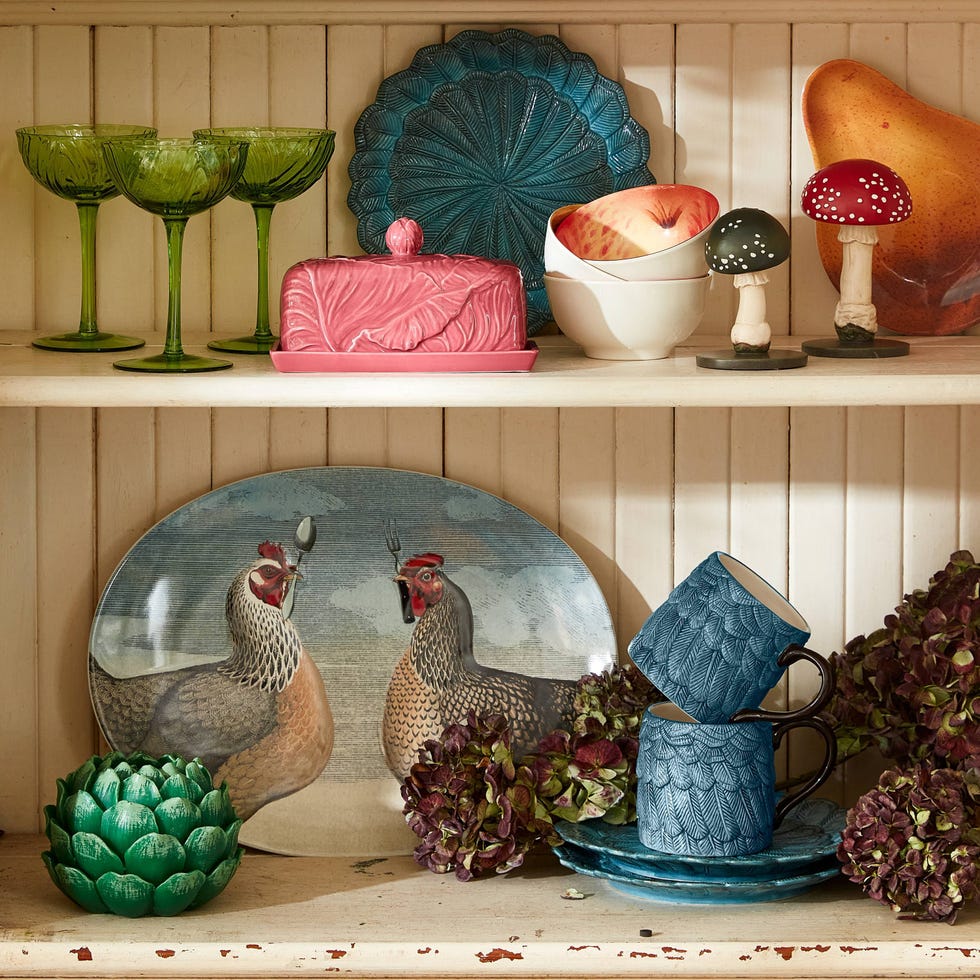 a shelving unit holds a variety of decorative items including green stemmed glasses a pink butter dish with a lid bowls with fruit and two ceramic mushroom figurines a prominent oval platter features illustrated chickens while textured blue mugs and plates are stacked nearby the colorful arrangement is accentuated by hydrangeas at the bottom