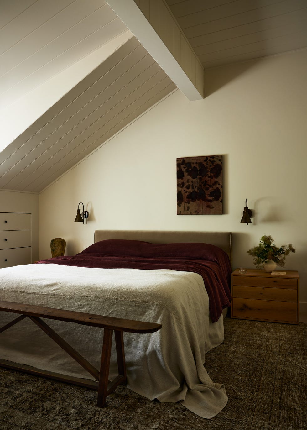 bedroom with white walls and slanted ceiling and a wooden bench at the end of a bed layered with linens