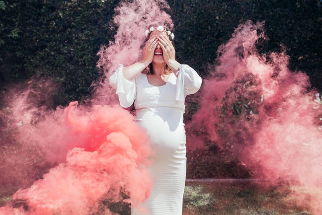 Happy pregnant woman covering her face with smoke grenade outdoors. Gender reveal party.