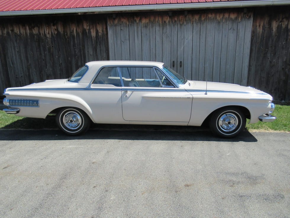 side view of a white 1962 dodge polara 500 parked on a paved driveway with a wooden barn in the background.
