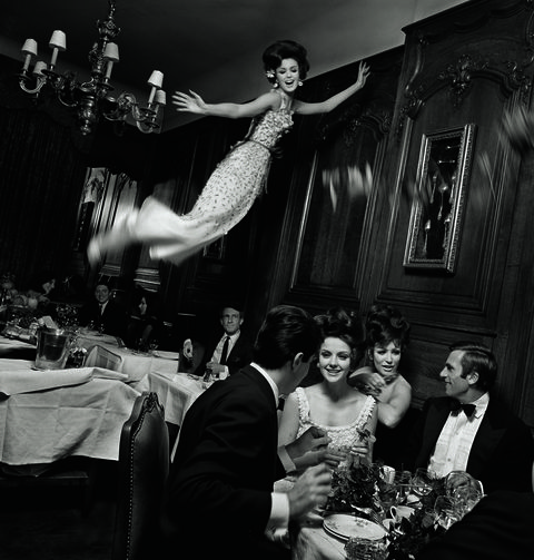 a model in an ornate evening dress towers over a dinner party in a formal room with a chandelier in this 1965 fashion photo