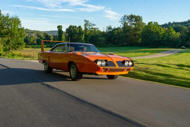 front three quarter view of orange coupe driving on rural road with trees and fields in background.