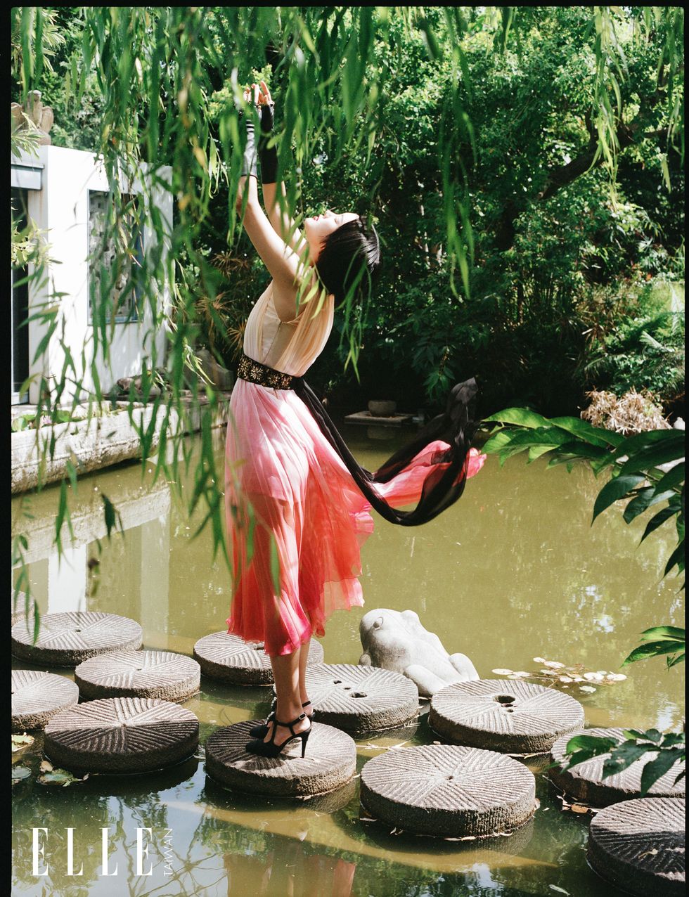 女人站在水中的藤墊上，身穿漸層百摺洋裝。