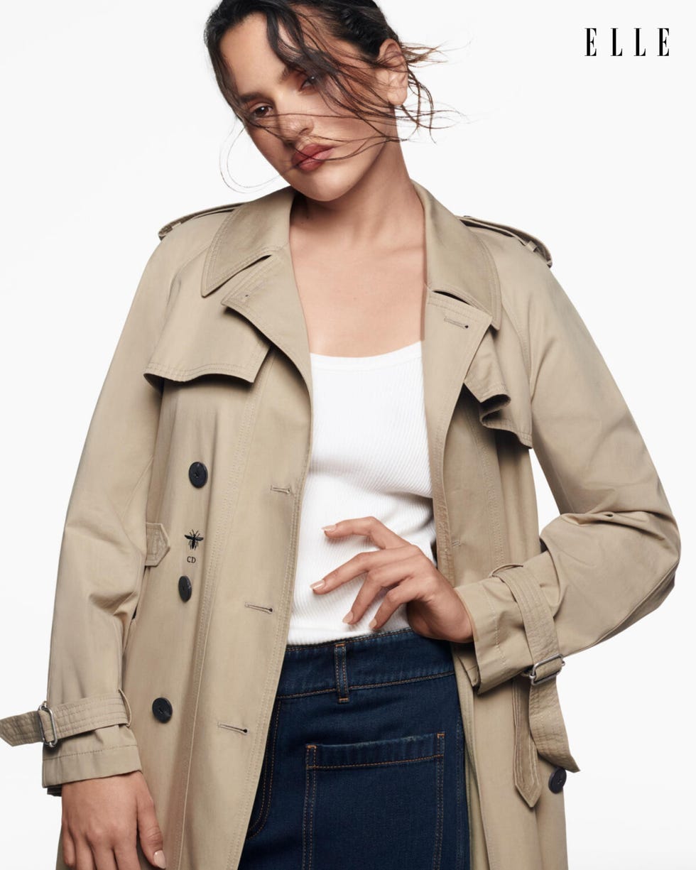 a person wearing a beige trench coat and a white tank top posing with a casual hand gesture