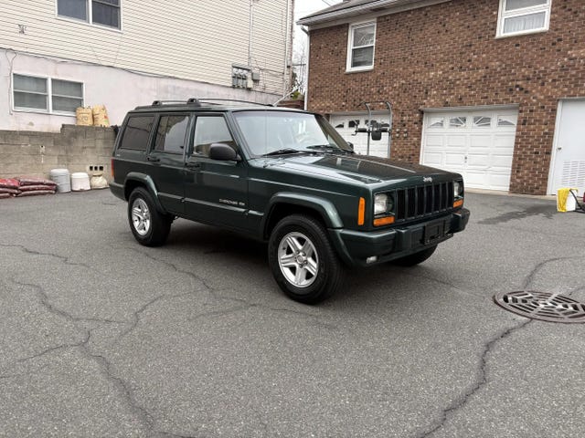front three quarter view of a green jeep cherokee 4x4 with a boxy design, silver alloy wheels, and orange turn signals.