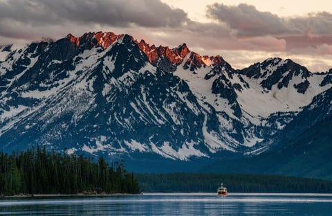 GRAND TETON NATIONAL PARK, WYOMING GRAND TETON NATIONAL PARK WYOMINGIn onze half leeggelopen kano peddelden we het water op alleen om de zonsondergang te bekijken maar door de bewolking leek het erop dat er die namiddag geen zonsondergang zou zijn Toen we weer naar de oever voeren draaide ik me om voor een laatste blik en zag dat het zonlicht net genoeg door de wolken scheen om de toppen van de bergen te beschijnen Keer om riep ik enthousiast en pakte haastig mn fototoestel Terwijl ik het juiste kader zocht dreef een bootje perfect in beeld Ik glimlachte en nam de foto
