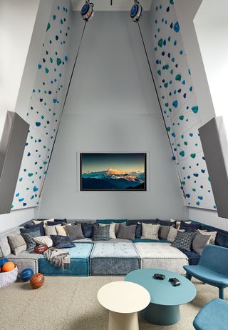 triangular shaped room with a high ceiling and climbing wall with blue holds on the side walls and a very large and very deep seating area composed of mattress like cushions and a million cushions all in shades of blue gray and cream and a small round pedestal table and armless chairs in front
