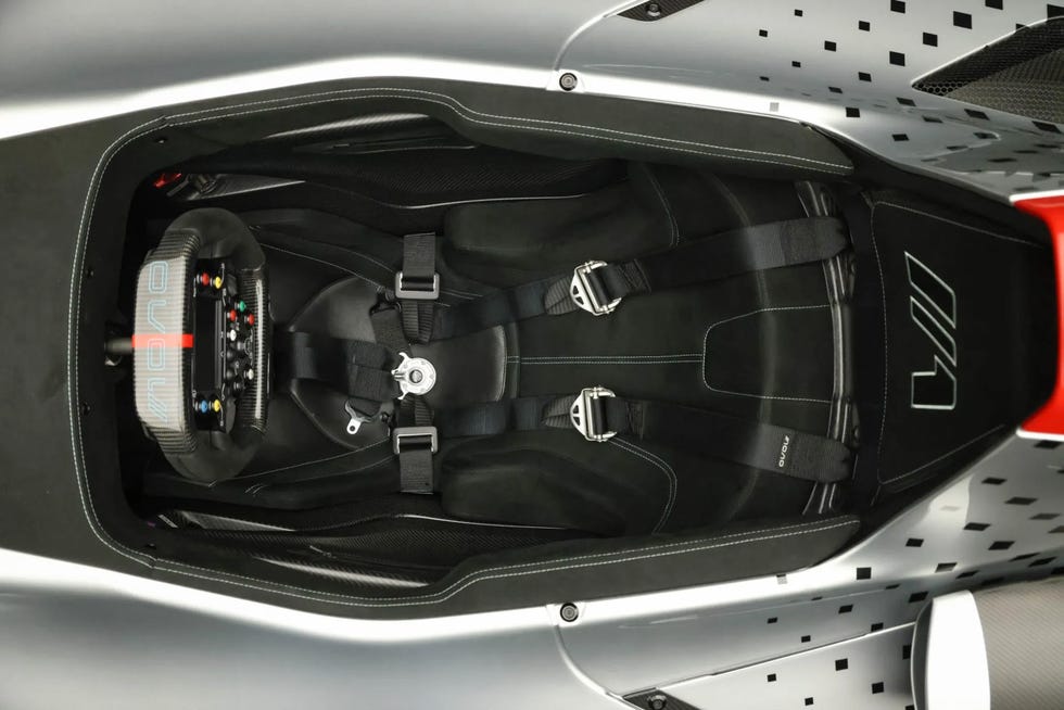 overhead view of a single seat cockpit with a racing harness and a carbon fiber steering wheel featuring integrated controls.