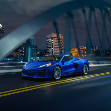 front 34 view of 2024 chevrolet corvette e ray 3lz coupe in riptide blue, driving over a bridge in front of a city pre production model shown actual production model may vary model year 2024 corvette e ray available 2023