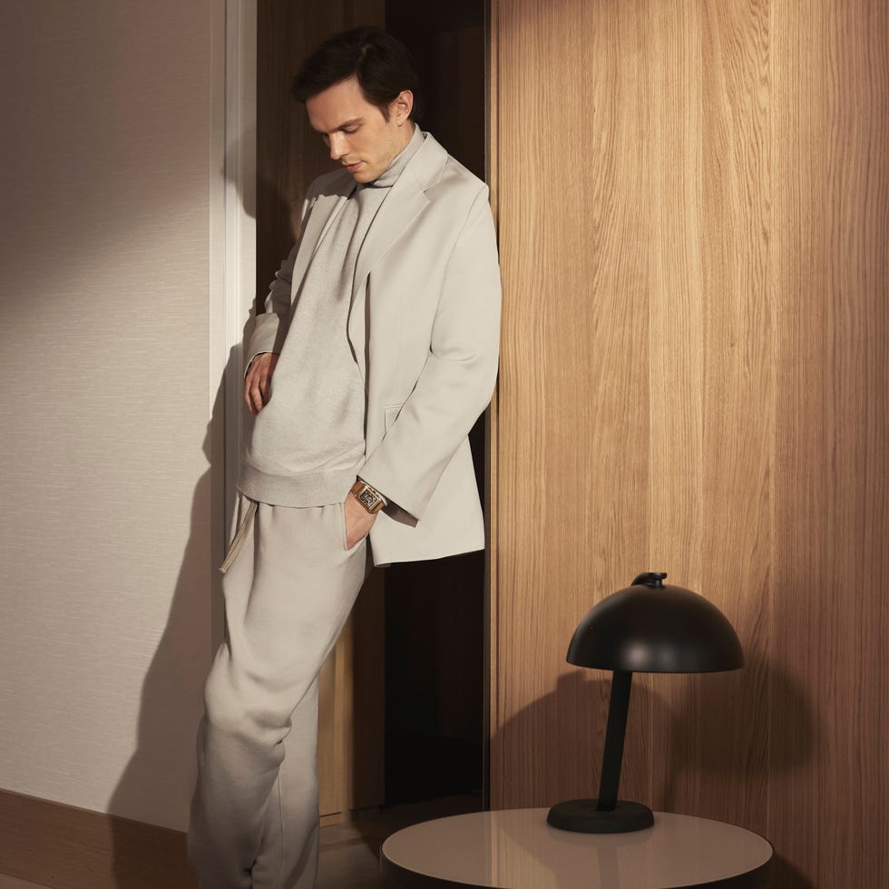 man in lightcolored outfit leaning against a doorway with a modern lamp nearby