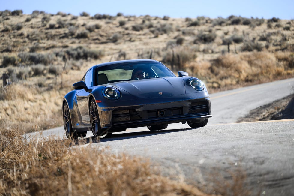 front view of a dark colored sports car driving on a winding road surrounded by dry grass and hills.