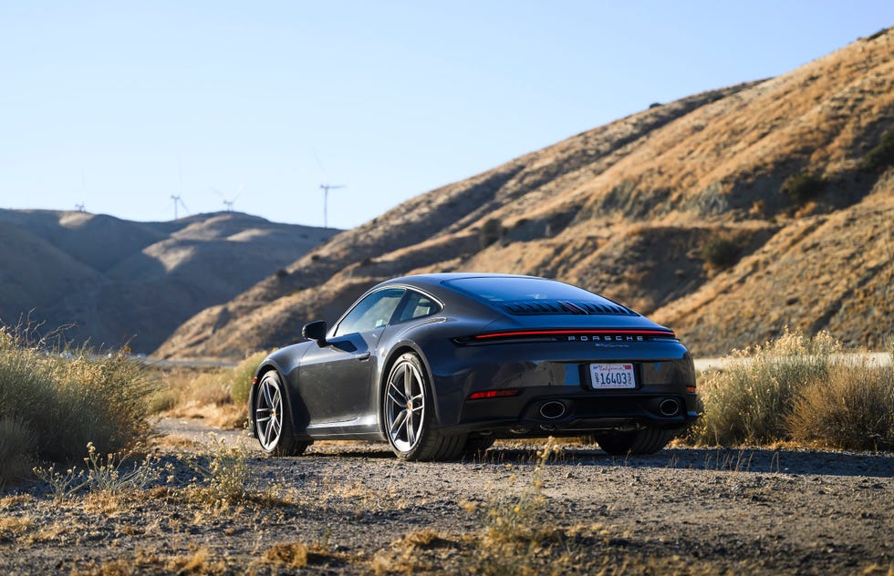 rear three quarter view of a dark colored sports car parked on a dirt path with hills and wind turbines in the background.