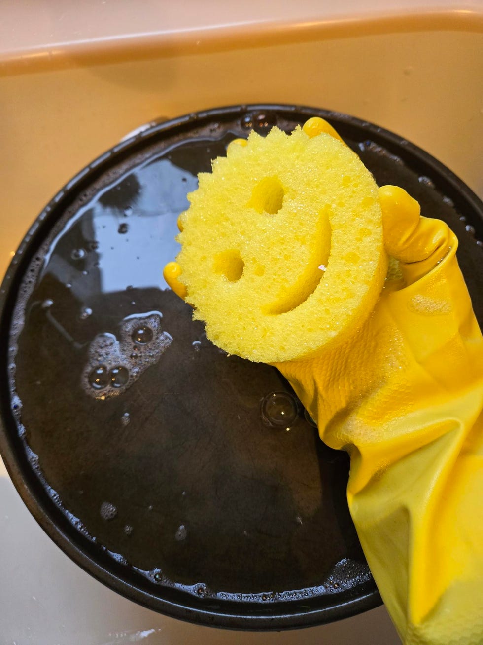 a smiling scrub daddy sponge held by a hand in a yellow glove over a soapy dish