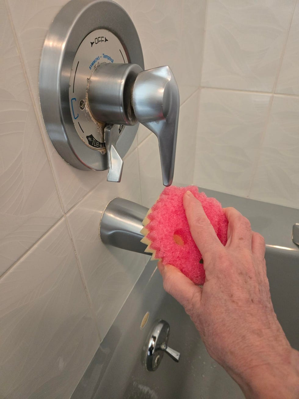 hand using a pink scrub mommy sponge to clean a bathtub faucet