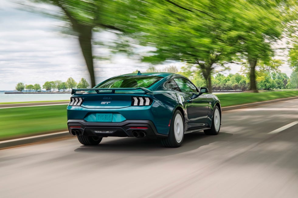 rear three quarter view of a blue sports car driving on a tree lined road near a body of water.