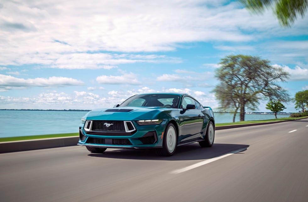 front three quarter view of a blue sports car driving along a lakeside road under a partly cloudy sky.