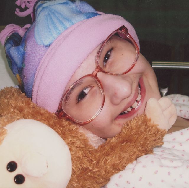 a girl wearing glasses and a pink and purple hat smiles as she lies in a bed next to a stuffed animal