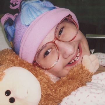 a girl wearing glasses and a pink and purple hat smiles as she lies in a bed next to a stuffed animal