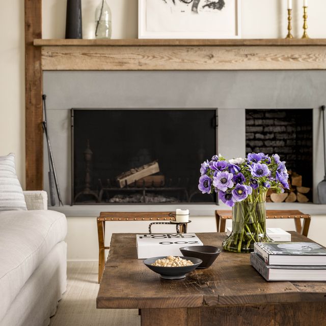 rustic room with wood beam mantle over a fireplace and log holder inset and a rough wood cocktail table and two modern stools and white sofa