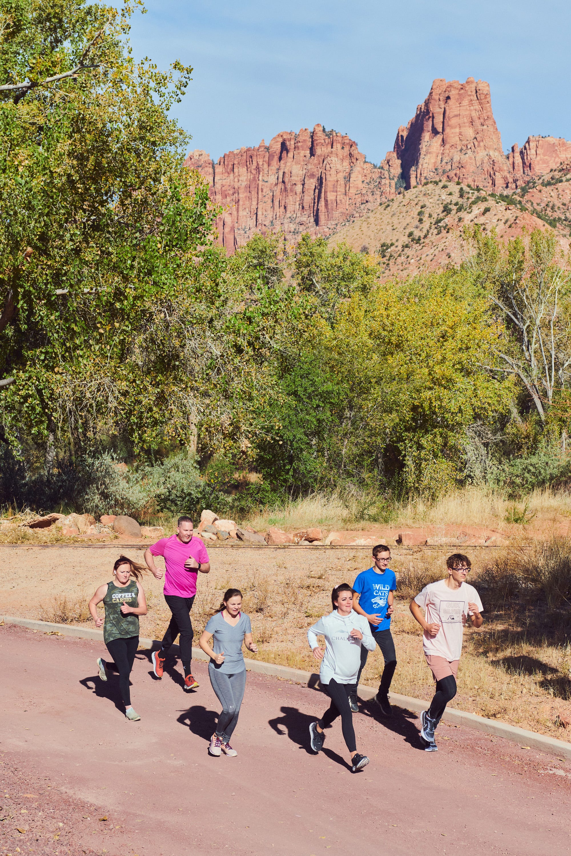 6 people run on a dirt road with a red rock formation in the background