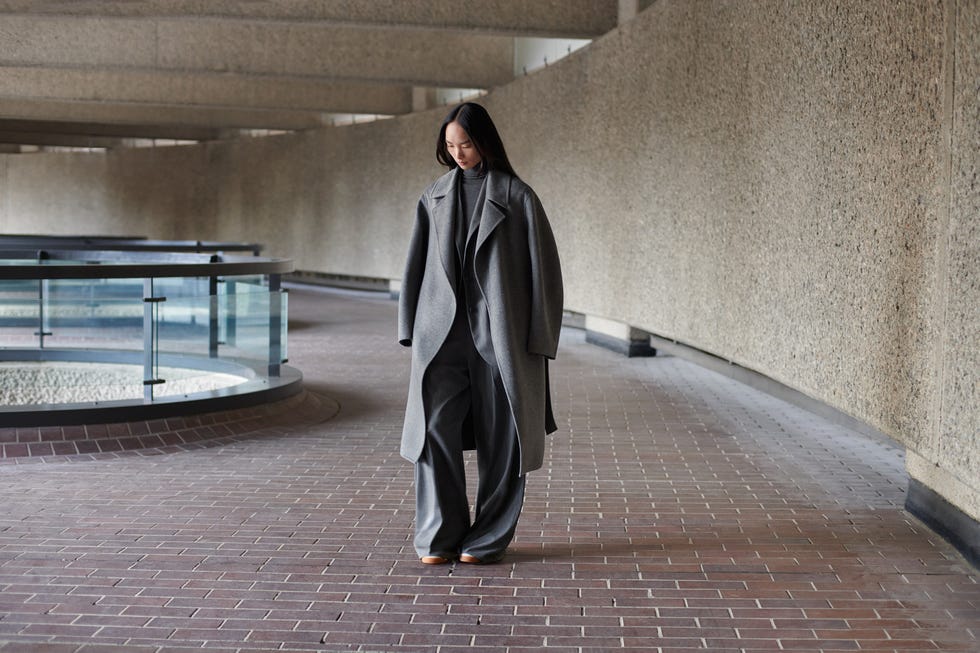 a person walking in a modern architectural space wearing a long grey coat and wide trousers