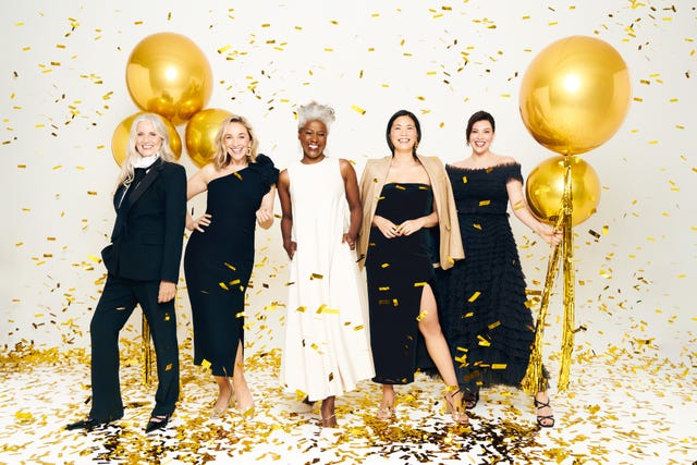 a group of five women stands together in a celebratory pose, surrounded by golden confetti falling from above they are dressed elegantly wearing various styles of black dresses and one white dress some hold large golden balloons, enhancing the festive atmosphere and the joyous occasion