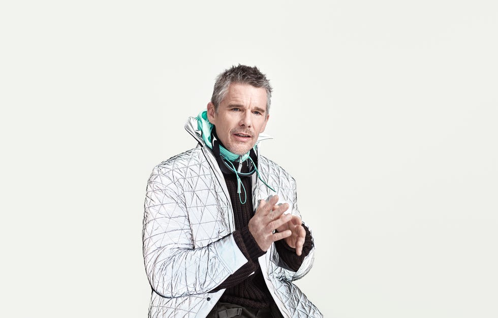 ethan hawke wearing a shiny quilted jacket and a hoodie sitting against a light background