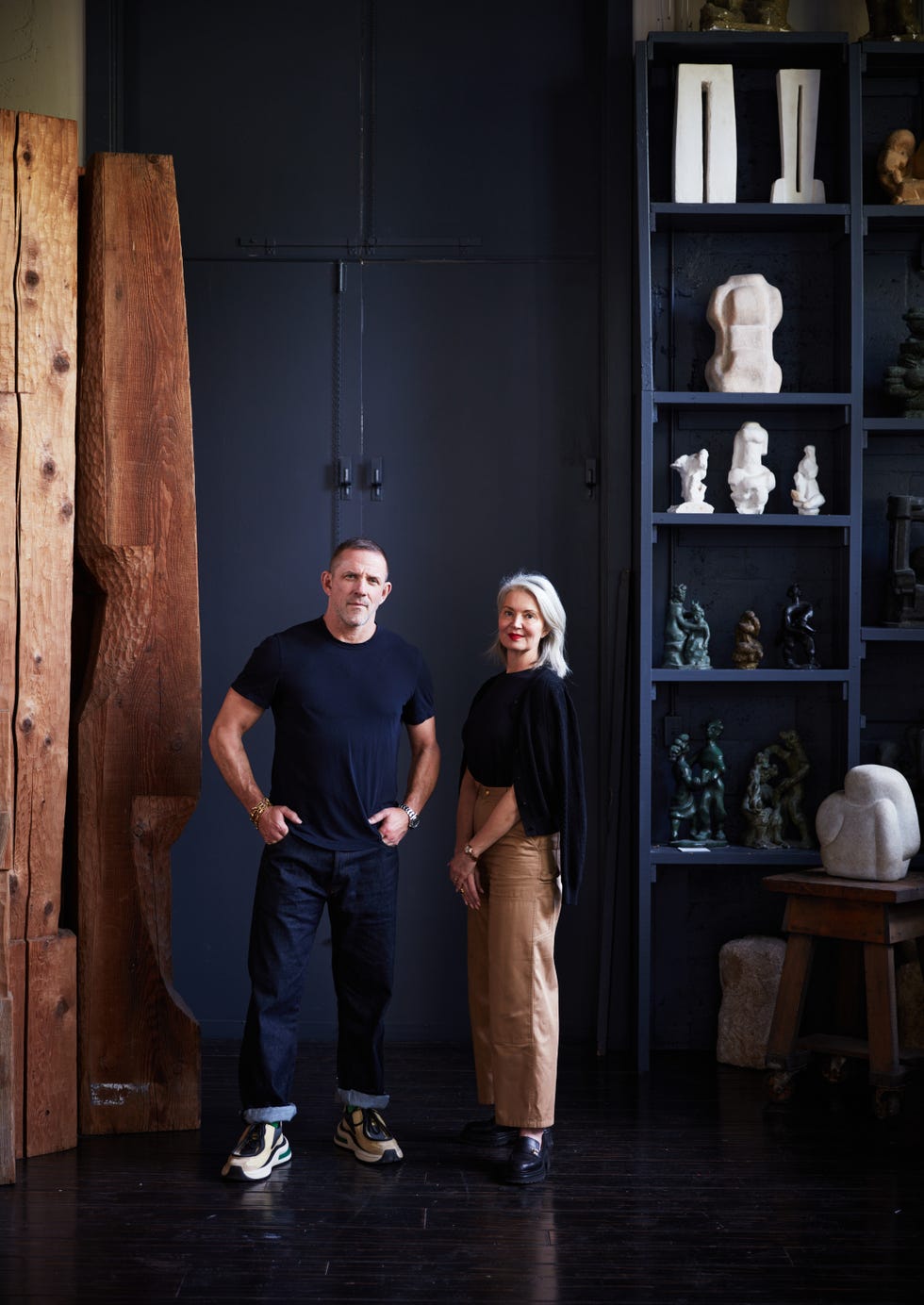 two individuals standing in a studio or gallery setting with sculptures and wooden elements