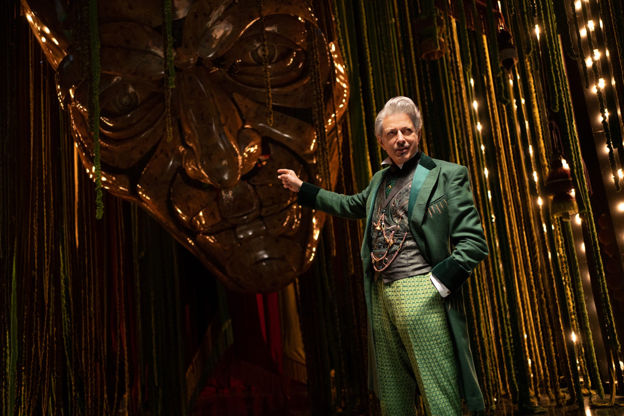 person in a green suit stands beside a large statue head