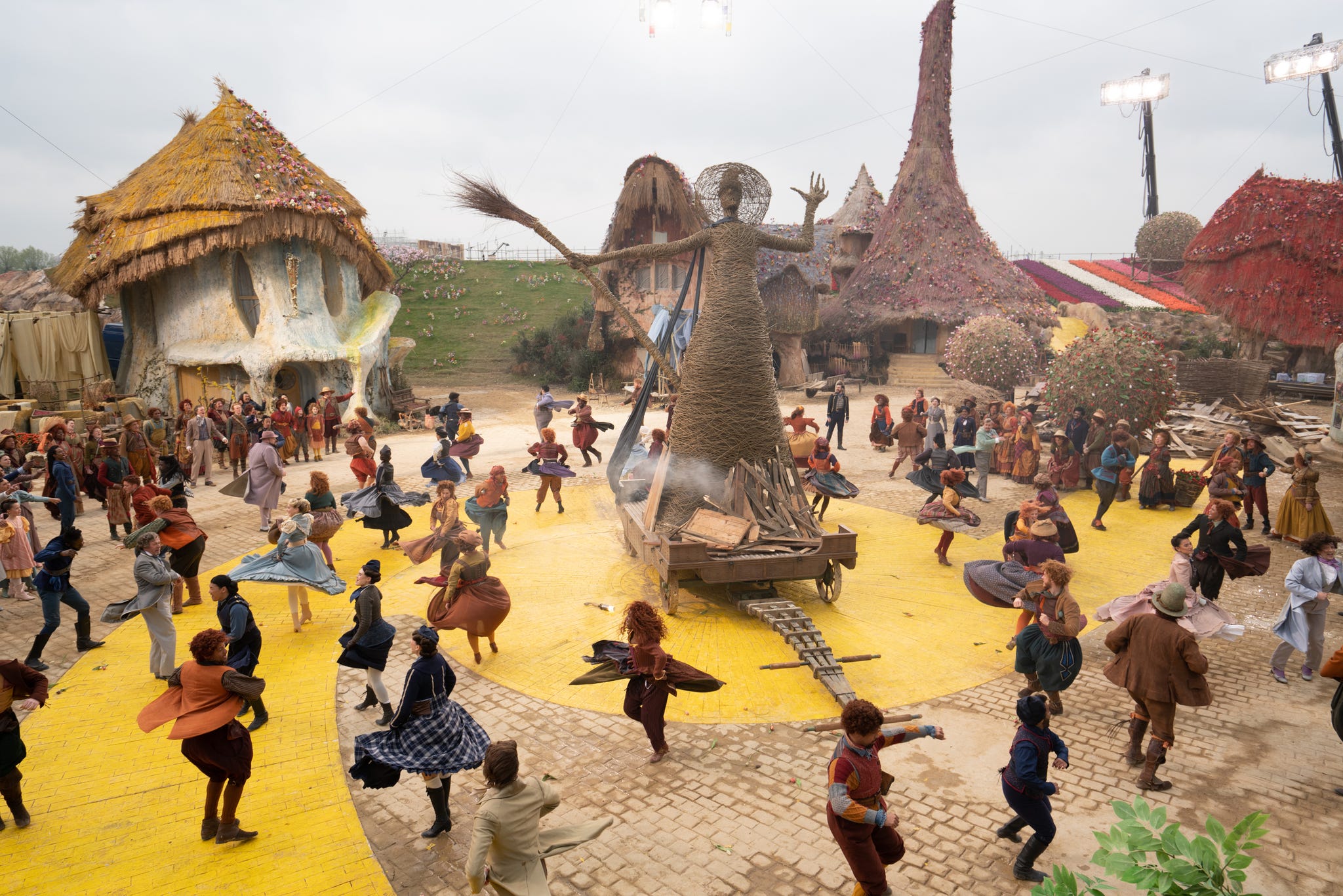 a lively dance scene in a village square with a large sculpture and several thatchedroof huts