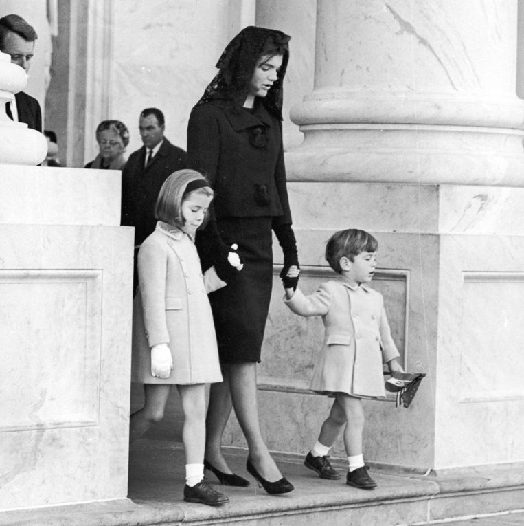 jacqueline kennedy holds the hands of her children caroline and john f kennedy jr as they walk down steps, jacqueline wears all black with a veil and the children wear light colored peacoats