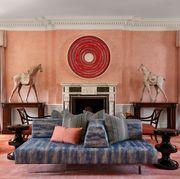 a pink walled and rugged living room with large red circular art over  white marble fireplace mantel with nothing on it and two tall wooden white saddled horses on three foot dark tables flanking the fireplace and facing it  as tall simple cream valanced curtain over french doors right next to each and in the center of the room is a large lounge sofa with blue stripped fabric and heaped with pillows