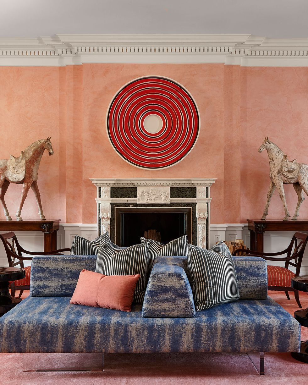 a pink walled and rugged living room with large red circular art over white marble fireplace mantel with nothing on it and two tall wooden white saddled horses on three foot dark tables flanking the fireplace and facing it as tall simple cream valanced curtain over french doors right next to each and in the center of the room is a large lounge sofa with blue stripped fabric and heaped with pillows