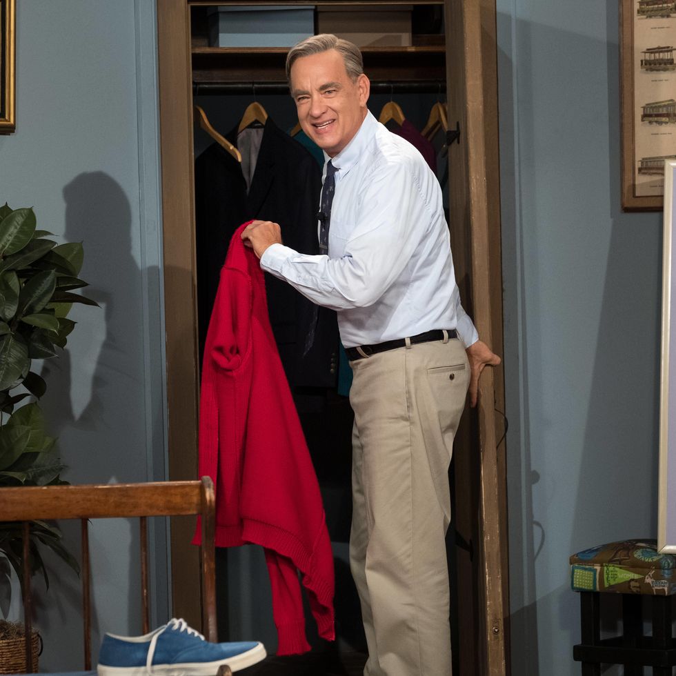tom hanks dress as fred rogers for a beautiful day in the neighborhood, he looks back at the camera and smiles while standing in front of a closet to hang a red jacket in his hand, he wears a white collared shirt, tie, and khaki pants