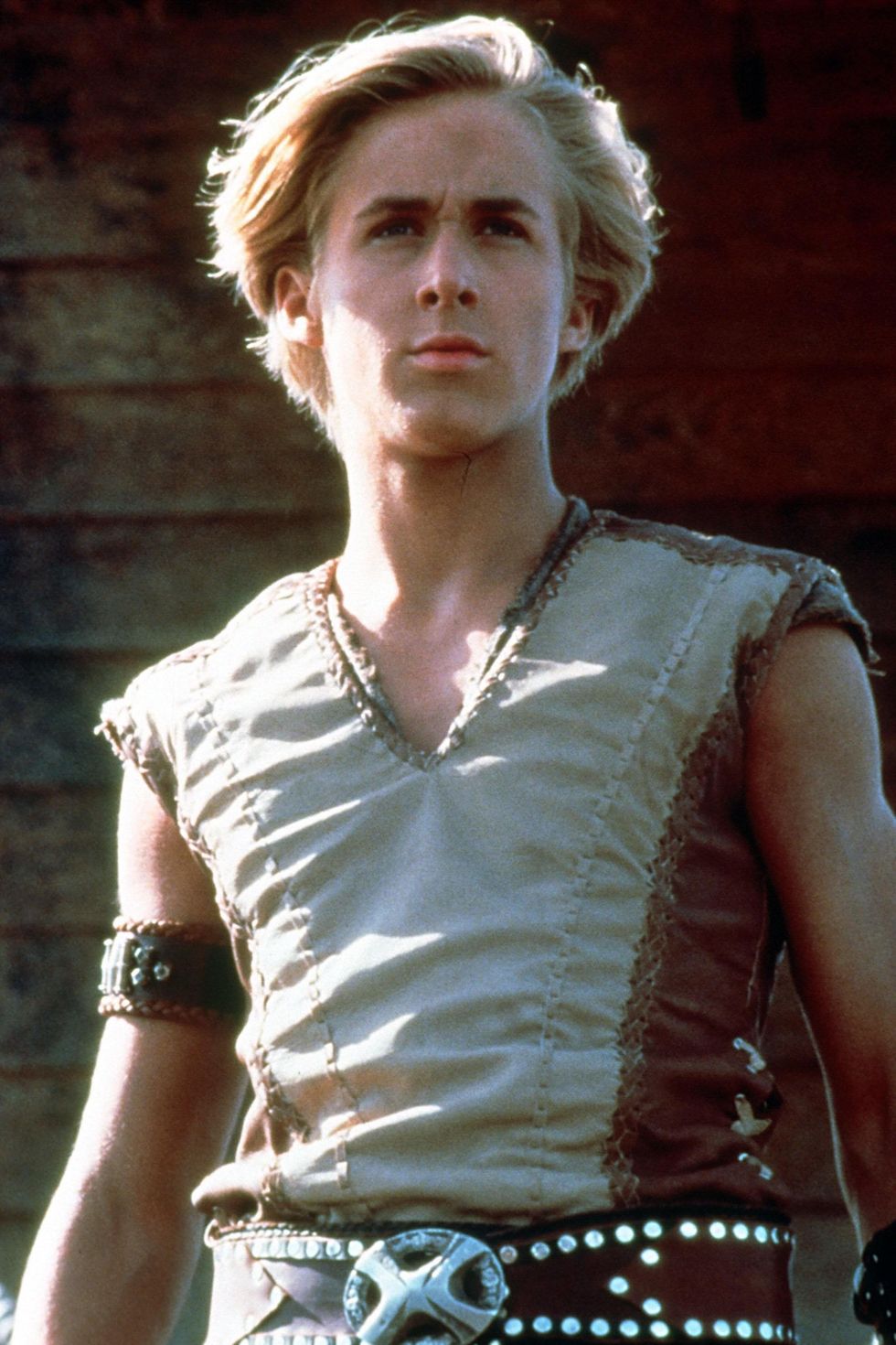 ryan gosling in character for young hercules, he wears a sleeveless tan top with an armband and a black belt