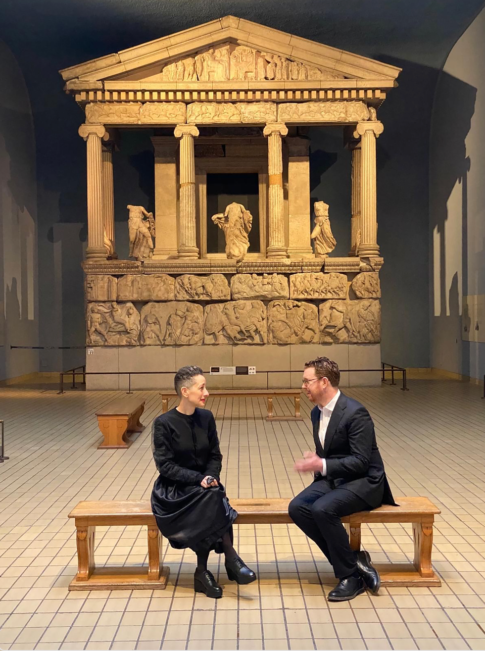 Two individuals engaged in conversation in front of a classical statue exhibit.
