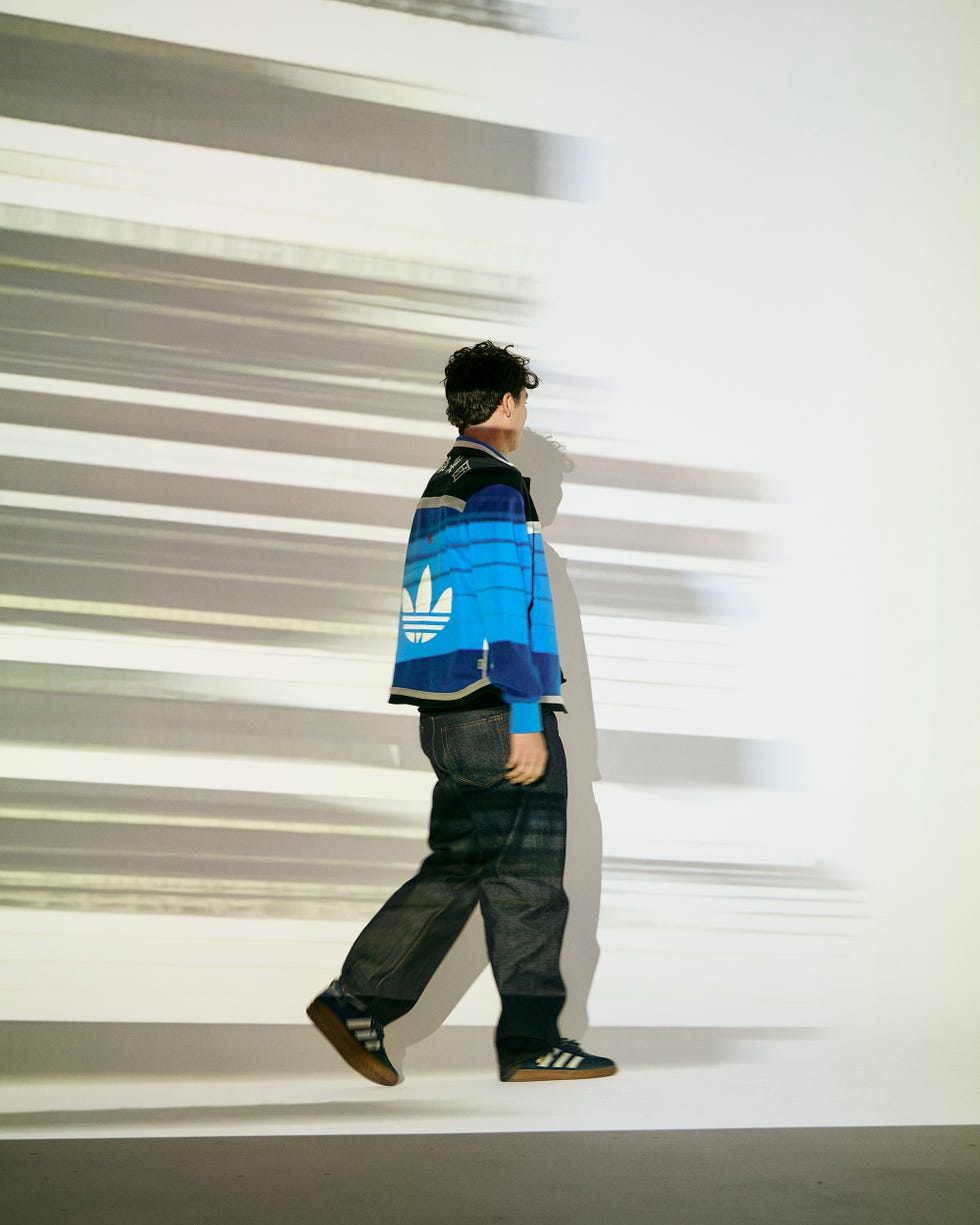 a person in a blue striped jacket and dark jeans walking in front of a light background with abstract lines