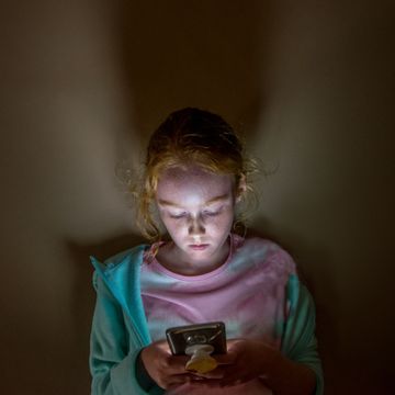 a young long haired redhead girl with face lit up looking into mobile phone screen in a dark room, against a flat wall, with sad facial expression