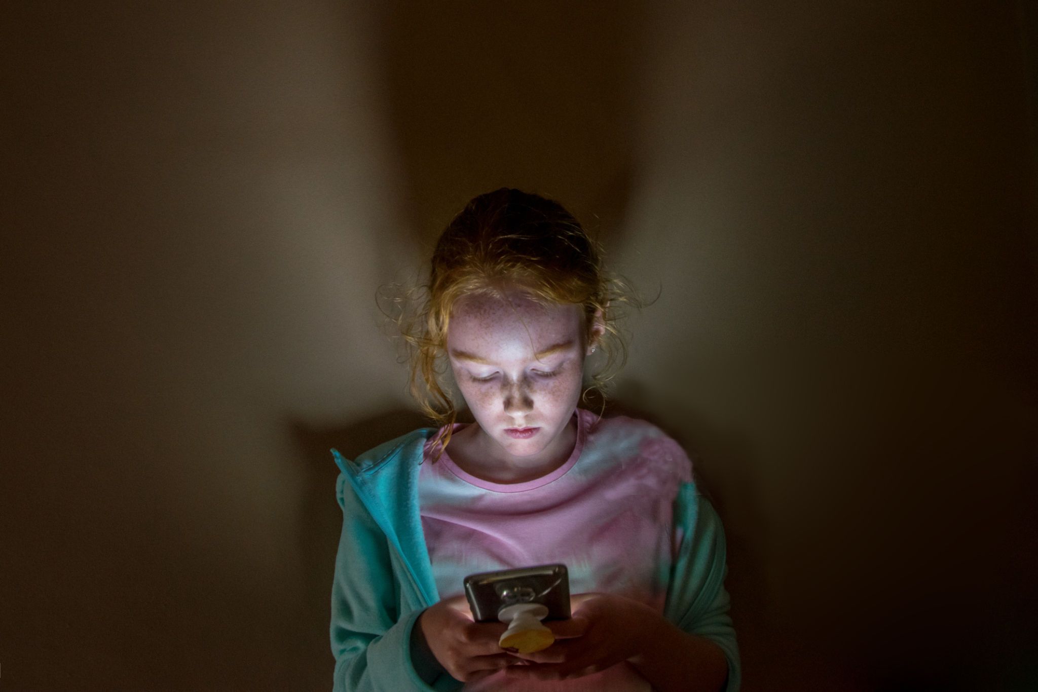 a young long haired redhead girl with face lit up looking into mobile phone screen in a dark room, against a flat wall, with sad facial expression