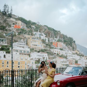 two woman sit on the bonnet of a vintage red convertible car enjoy touring italy together