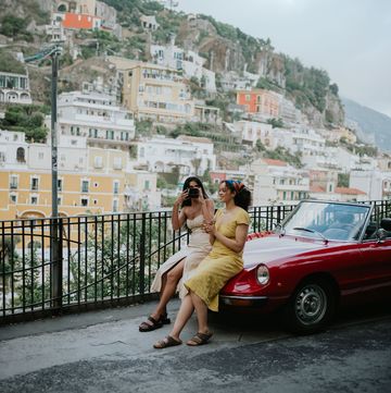 two woman sit on the bonnet of a vintage red convertible car enjoy touring italy together