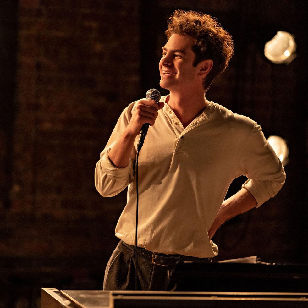 andrew garfield as jonathan larson for tick, tick, boom, he wears a cream colored long sleeve t shirt with brown pants and holds a microphone while smiling and looking left