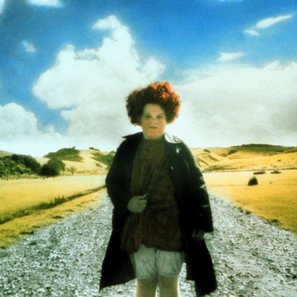 alexia keogh in character as janet frame for an angel at my table, she stands on a gravel road wearing a jacket, blouse and shorts