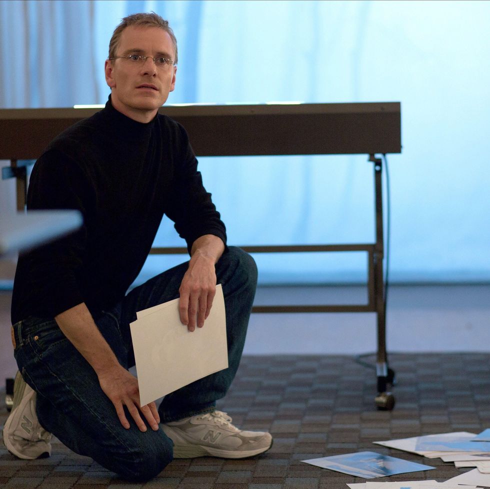 michael fassbender dressed as steve jobs crouches on a rug while holding a piece of paper in his hand and looking toward the camera, he has on a black turtleneck and black pants with white sneakers and wire rimmed glasses