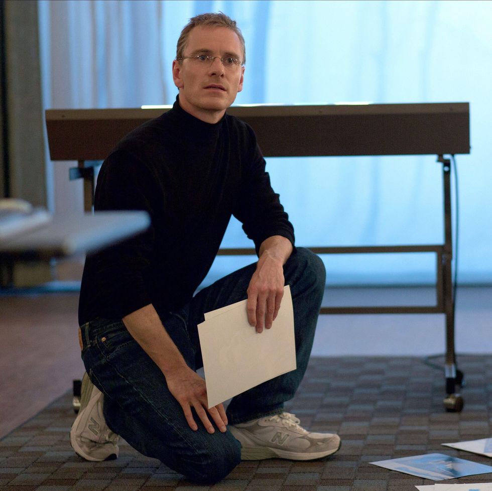 michael fassbender dressed as steve jobs crouches on a rug while holding a piece of paper in his hand and looking toward the camera, he has on a black turtleneck and black pants with white sneakers and wire rimmed glasses