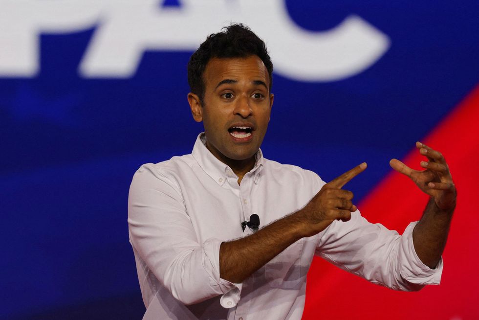 vivek ramaswamy speaks at the conservative political action conference in dallas, texas, on august 5, 2022, he wears a white button up shirt with a collar and has a microphone clipped to the front of his shirt, both of his arms are raised out in front of him and his right hand points to his left hand that is in an open c shape, behind him is a blue and red background with the letters cpac