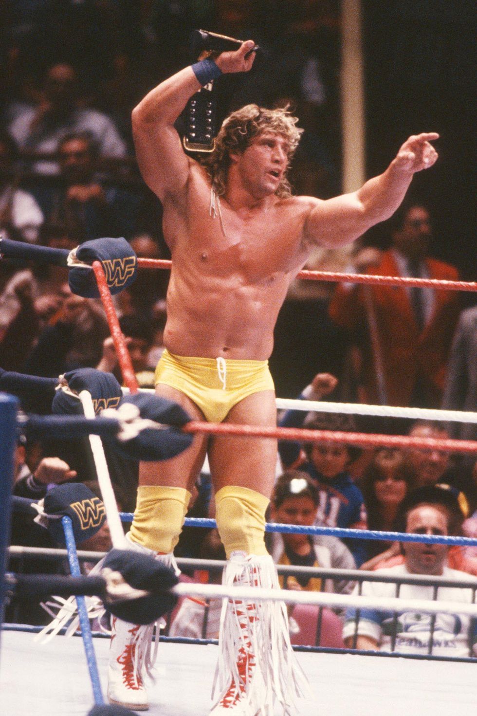 kerry von erich stands inside a wrestling ring holding a belt over his head with one hand and pointing right with his other, he wears yellow shorts and knee pads with knee high white boots with long fringe and red laces