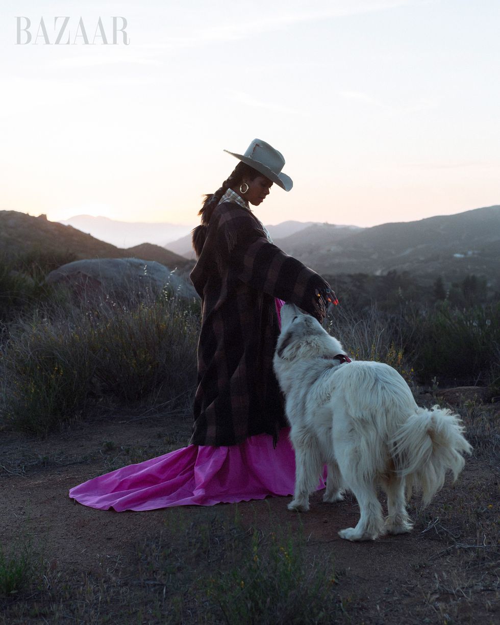 kelis wears cowboy hat, long coat, and pink dress standing next to a dog on a hill