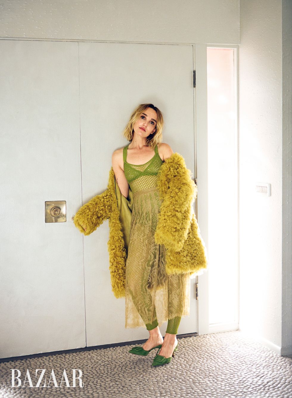 chloe fineman leans against a white door wearing a green knit bodysuit, sheer lace skirt, green mules, and a yellow shag jacket